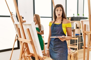 Wall Mural - Young hispanic woman at art classroom scared and amazed with open mouth for surprise, disbelief face