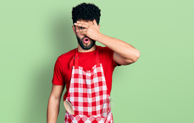 Sticker - Young arab man with beard wearing cook apron peeking in shock covering face and eyes with hand, looking through fingers with embarrassed expression.