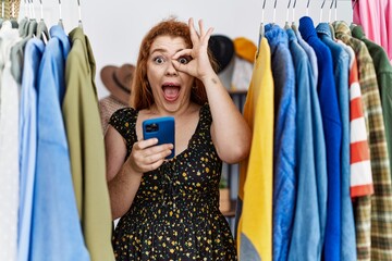 Sticker - Young redhead woman searching clothes on clothing rack using smartphone smiling happy doing ok sign with hand on eye looking through fingers