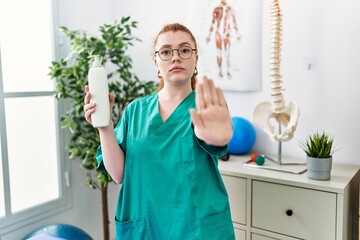 Canvas Print - Young redhead physiotherapist woman holding massage body lotion with open hand doing stop sign with serious and confident expression, defense gesture