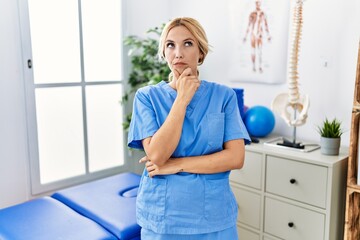 Canvas Print - Beautiful blonde physiotherapist woman working at pain recovery clinic looking confident at the camera with smile with crossed arms and hand raised on chin. thinking positive.