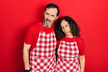 Wall Mural - Middle age couple of hispanic woman and man wearing cook apron in shock face, looking skeptical and sarcastic, surprised with open mouth