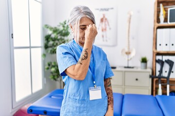 Poster - Middle age grey-haired woman wearing physiotherapist uniform at medical clinic tired rubbing nose and eyes feeling fatigue and headache. stress and frustration concept.