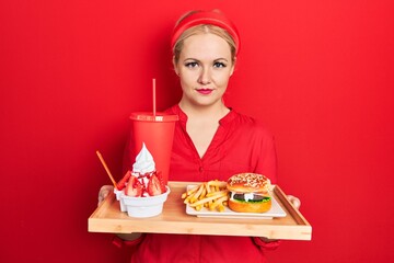 Canvas Print - Young blonde woman eating a tasty classic burger with fries and soda relaxed with serious expression on face. simple and natural looking at the camera.