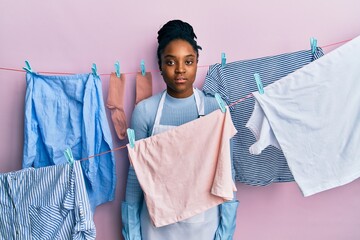 Poster - African american woman with braided hair washing clothes at clothesline relaxed with serious expression on face. simple and natural looking at the camera.