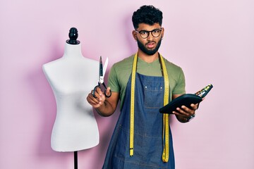 Poster - Arab man with beard dressmaker designer holding scissors and sewing kit in shock face, looking skeptical and sarcastic, surprised with open mouth