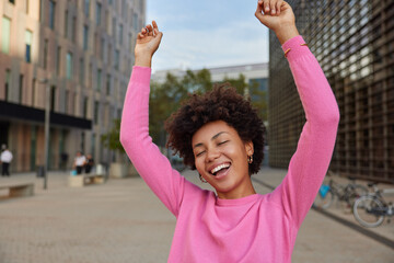 Carefree positive woman with curly hair keeps arms raised up dances with joy walks at city street wears casual pink jumper keeps eyes closed from happiness. Freedom relax and happiness concept