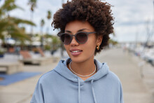 Outdoor Shot Of Pleased Woman With Curly Hair Wears Sunglasses And Hoodie Walks At Pier Harbor Admires Scenic Views. Female Tourist On Shore Strolls During Daytime. Tourism And Recreation Concept