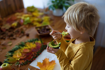 Wall Mural - Sweet child, boy, eating avocado while applying leaves using glue while doing arts and chraft with leaves