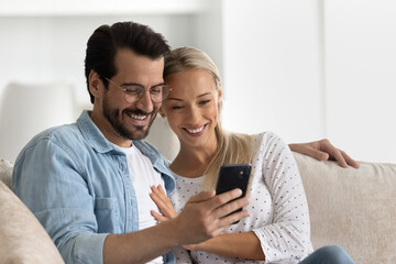 Wall Mural - Happy bonding loving married family couple using cellphone, resting on cozy sofa at home. Affectionate young man in eyeglasses cuddling smiling wife, web surfing information or playing games.