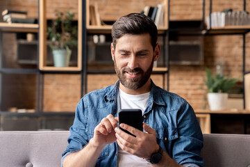 Smiling man using mobile phone scrolling network feed sitting on sofa at home 