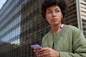 Wall Mural - Outdoor shot of pensive young curly female model uses smartphone for blogging chats online connected to wireless internet types content text for sharing for web dressed in jacket looks away.