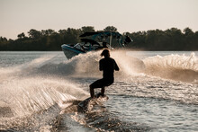 Rear View Of Active Man Riding Wakeboard Behind Motor Boat On Splashing River Waves. Active And Extreme Sports