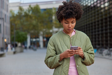 Wall Mural - Outdoor shot of young curly woman uses modern smartphone for chatting checks income mail shares media wears jacket walks against blurred city background. People technology and free time concept