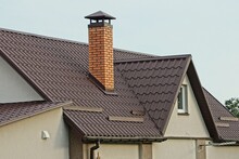 One Large Red Brick Chimney On A Brown Tiled Roof Of A Private Gray House With A Window Against The Sky