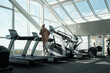 © pressmaster - Back view of young active female running on treadmill in front of large window in contemporary leisure center