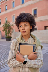 Wall Mural - Vertical shot of beautiful curly haired woman carries notepad and tablet with app for students wears checkered shirt and vest walks oudoors against city building. Technology and lifestyle concept