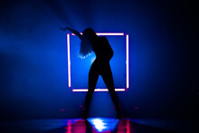 Silhouette Of Sexy Woman Dancing On Glowing Square Of Led Lamps Background. She Looks Seductively. Sexy Outfit . Blue Smoky Studio.