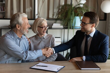 Shaking Hands. Smiling Young Man Real Estate Broker Handshaking With Older Family Couple After Buying Selling Property. Spouses Retirees Thank Bank Agent For Help After Signing Loan Mortgage Agreement