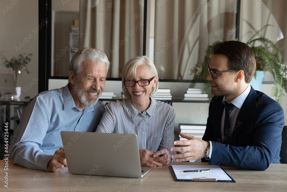 Smiling senior couple sit at lawyer broker designer office look at laptop listen to offer proposal of qualified specialist. Happy older spouses customers of male realtor choose flat house on pc screen