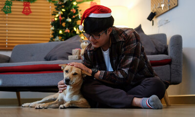 Young asian man wearing Santa hat sitting on floor with his dog in living room.