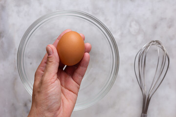 Wall Mural - Top view of left woman hand holding one raw brown egg above glass bowl and whisk on marble surface