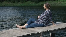 Handheld Shoot Pregnant Woman Sitting On Wooden Pier Near Lake And Stroking Her Belly. Healthy Pregnancy, Wellbeing Concept.