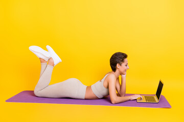 Poster - Profile side view portrait of attractive slender cheerful girl lying on mat using laptop isolated over bright yellow color background