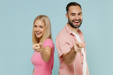 Side view young smiling fun couple two friends family man woman in casual clothes stand back to back point finger camera on you together isolated on pastel plain light blue background studio portrait.