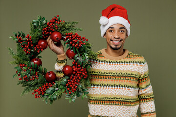 Young smiling happy african man in knitted sweater red Christmas Santa Claus hat hold Advent wreath isolated on plain green khaki background studio portrait Happy New Year 2022 celebration concept.