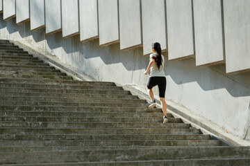 Slim woman with flying hair in tracksuit runs up large stone steps past wall with concrete plates on city street backside view