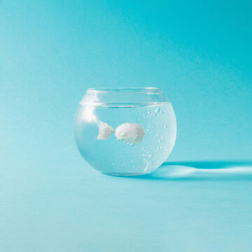 Creative composition with fish bowl and candy wraped in white plastic against blue background. Minimal ecology concept.