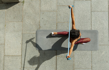 Young biracial sportswoman in elegant tracksuit does dynamic lunges holding elastic band standing on sunny pavement upper view