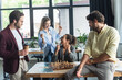 © LIGHTFIELD STUDIOS - Interracial businesswomen showing yes gesture near colleagues playing chess in office