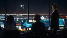 Female And Male Air Traffic Controllers With Headsets Talk In Airport Tower At Night. Office Room Full Of Desktop Computer Displays With Navigation Screens, Airplane Flight Radar Data For Controllers.
