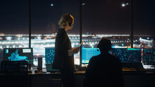 Female And Male Air Traffic Controllers With Headsets Talk In Airport Tower At Night. Office Room Full Of Desktop Computer Displays With Navigation Screens, Airplane Flight Radar Data For Controllers.