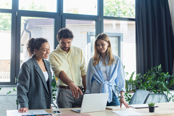 Wall Mural - Businessman pointing at laptop near smiling interracial businesswomen in office