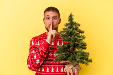 Young caucasian man buying little tree for Christmas  isolated on yellow background keeping a secret or asking for silence.