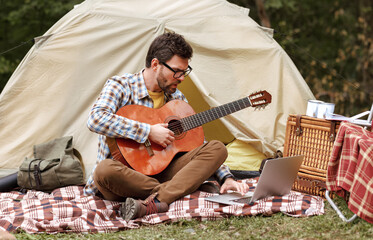 Young man learning to play acoustic guitar through internet on laptop during camping in forest