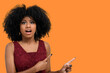 © Edson Souza - Positive young woman with afro hairdo, points to the side with a cheerful expression, shows something incredible in the blank space, isolated on a orange background