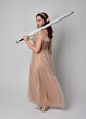 © faestock - Full length  portrait of red haired  girl wearing a creamy fantasy gown and crystal crown, like a fairy goddess costume.  Standing pose holding a sword weapon  isolated on light studio background.