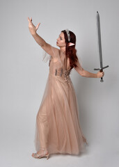 Full length  portrait of red haired  girl wearing a creamy fantasy gown and crystal crown, like a fairy goddess costume.  Standing pose holding a sword weapon  isolated on light studio background.