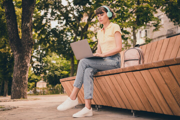 Full body photo of cute millennial brunette lady sit work laptop wear polo headphones jeans sneakers outside in city