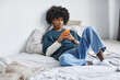 © Seventyfour - Minimal full length portrait of young African-American man using smartphone on bed at home, copy space