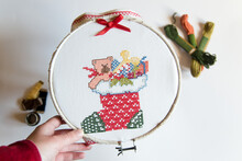 Bastidor De Bordado Con Un Trozo De Tela Con Un Motivo Navideño Realizado En Punto De Cruz. Diseño De Un Calcetín Lleno De Regalos. Lazo Rojo.