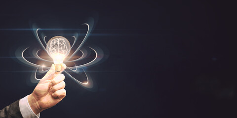 close up of hand holding abstract light bulb on dark wide background with digital brain and atom out