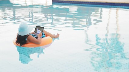 Wall Mural - woman in a sun hat and a swimsuit in the pool in a swimming circle with a smartphone drinks a cocktail with her back to the camera. Overall plan