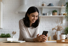 Happy Joyful Young Asian Woman Reading Text Message On Mobile Phone Screen, Chatting Online, Watching Funny Video, Media Content. Smiling Gen Z Student Girl Using Smartphone In Home Kitchen, Laughing