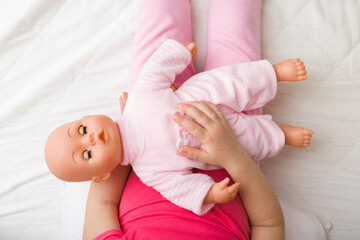 Toddler girl sitting on bed, holding and putting to sleep baby doll on lap. Point of view shot. Closeup. Child playing mother. Top down view.