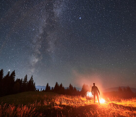 Wall Mural - silhouette of male tourist standing near campfire under beautiful night sky with stars and milky way
